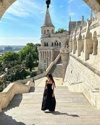 A woman in a black dress stands on the steps of a castle, looking out at the city below.