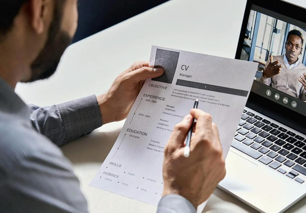 A man is holding a resume and looking at a laptop with a video call with another man.